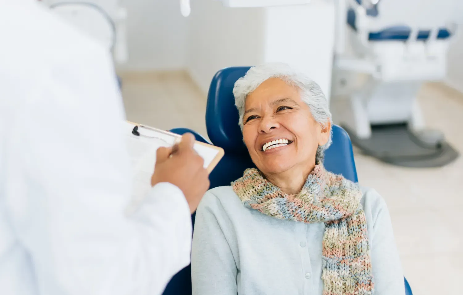  An older woman wearing a scarf smiles while seated in a dental chair, speaking with a dentist holding a clipboard.